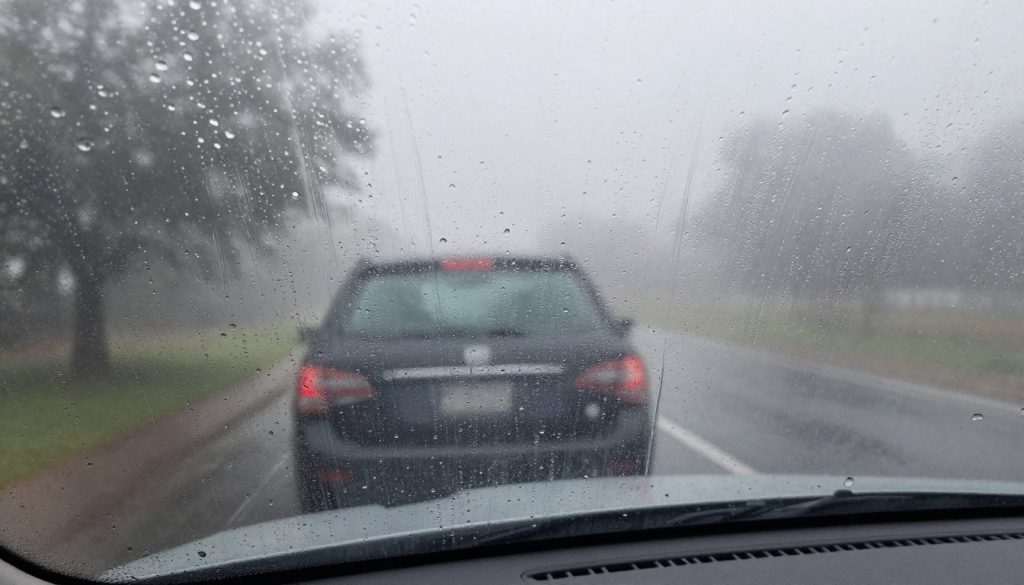 A close-up view of a car windshield on a foggy day, showcasing intricate patterns of condensation forming on the glass surface. In the foreground, focus on droplets merging and trickling down, catching the light in a soft, diffused way. The middle layer features a slightly blurred interior view of a car, hinting at a passenger seat and a dashboard, evoking a sense of being inside the vehicle. The background reveals a gloomy, humid Texas atmosphere with misty trees visible through the foggy glass, creating a tranquil yet slightly melancholic mood. The lighting is natural and soft, mimicking overcast conditions, with a shallow depth of field to emphasize the condensation on the windshield.